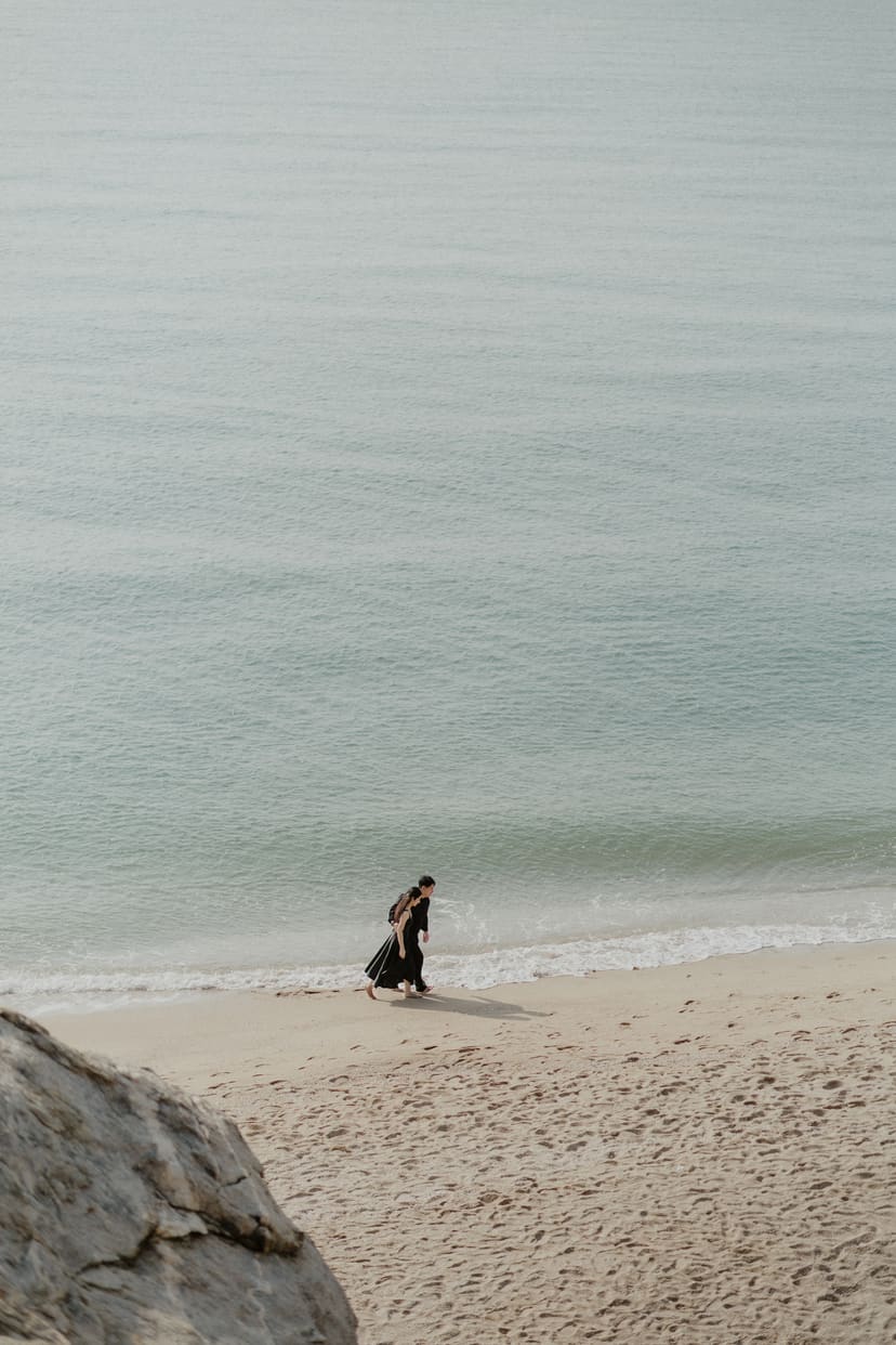The couple, small in the frame, standing at the edge of a grey-green sea on an overcast day